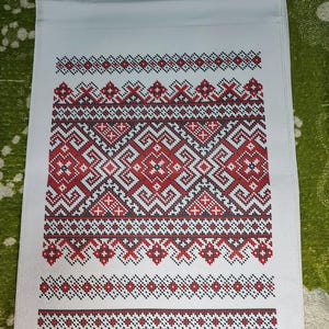 May include: A white rectangular fabric runner featuring a central panel of intricate cross-stitch embroidery. The pattern consists of repeating geometric shapes and linear borders in vibrant red and black thread against the plain white background.
