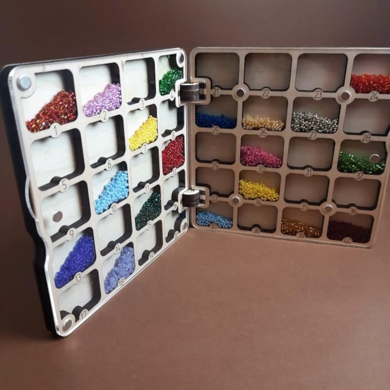 K&ouml;nnte beinhalten: Ein offener Holzorganizer mit zahlreichen F&auml;chern, die jeweils mit farbigen Perlen gef&uuml;llt sind. Der Organizer zeigt eine Vielzahl von Perlenfarben, darunter Rot, Lila, Gelb, Blau und Gr&uuml;n. Die F&auml;cher sind mit Nummern versehen.