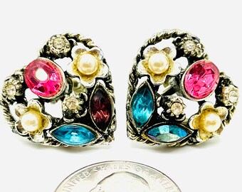 Op de afbeelding: Een paar hartvormige oorbellen met een zilverkleurig metalen frame. Elke oorbel heeft een grote roze ovale edelsteen, een blauwe marquise-geslepen edelsteen en een kleinere paarse edelsteen. Accentueren met kleine heldere kristallen, imitatieparels en bloemdetails.