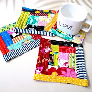 May include: A white mug with the word "LOVE" printed on it rests on a colourful patchwork coaster. The coasters are square, with a variety of fabrics in red, blue, green, and yellow. The coasters are approximately 10 cm square.