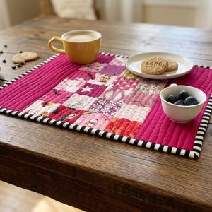 May include: A patchwork placemat in pink, red, and purple, with a black and white striped border. A yellow mug, a plate with biscuits, and a small bowl of blueberries are on the placemat. The placemat measures approximately 30 cm x 40 cm.