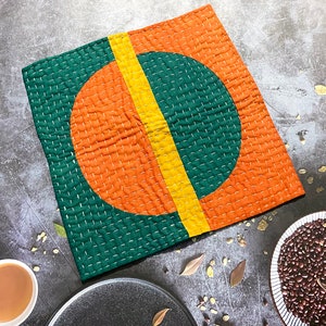May include: A green, orange and yellow quilted fabric square with a stitched circle design. The circle is divided in half, with one half orange and the other half green. A yellow line runs down the centre of the circle.