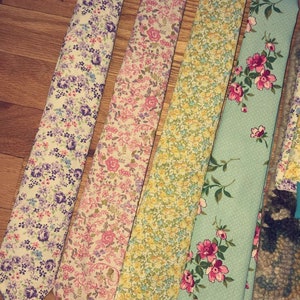 May include: Four neckties arranged side by side, each featuring a distinct floral pattern. The first has purple and blue flowers on white, the second pink and green florals, the third yellow and green blossoms, and the fourth pink flowers on light blue.