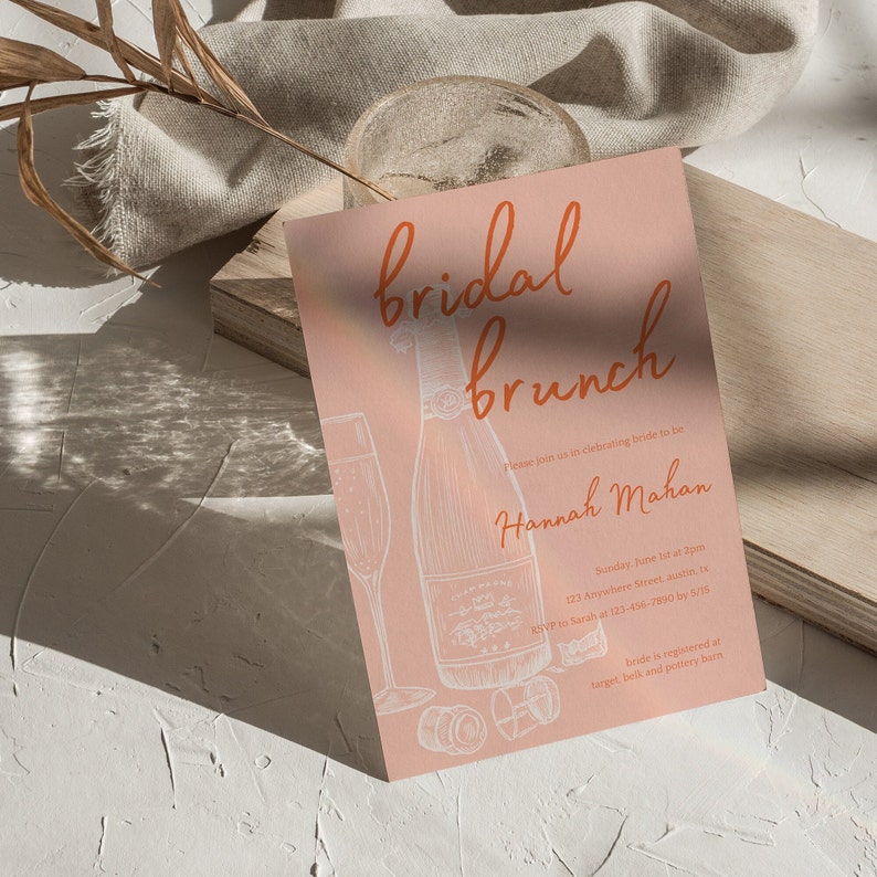 May include: A peach-colored bridal brunch invitation with a white illustration of a champagne bottle and glass. The text reads "bridal brunch" in orange cursive lettering and "Please join us in celebrating bride to be Hannah Mahan" in orange lettering. The invitation includes details for the event, including the date, time, location, and RSVP information.