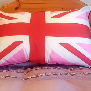 Pode incluir: Uma almofada rosa e vermelha com um design Union Jack. A almofada está em uma cama com uma colcha de cor roxa clara com babados.