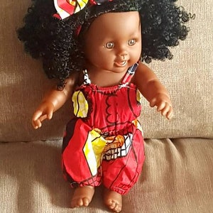 Könnte beinhalten: Eine schwarze Puppe mit lockigem Haar, die einen rot-gelb gemusterten Strampler und ein rot-gelb-blaues Stirnband trägt.