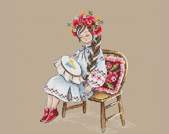 May include: A woman in a blue and white embroidered dress sits on a wooden chair with a floral cushion. She wears a wreath of red flowers and is holding a hoop with blue and yellow embroidery.