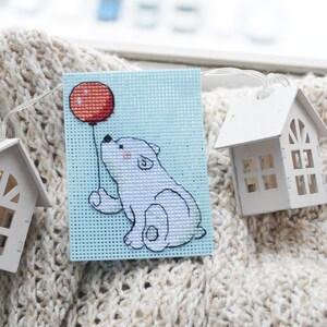 May include: A cross-stitch art piece featuring a white polar bear holding a red balloon against a light blue background. The bear has pink cheeks and is stitched on a square of perforated canvas. Two small white house-shaped decorations are in the background.