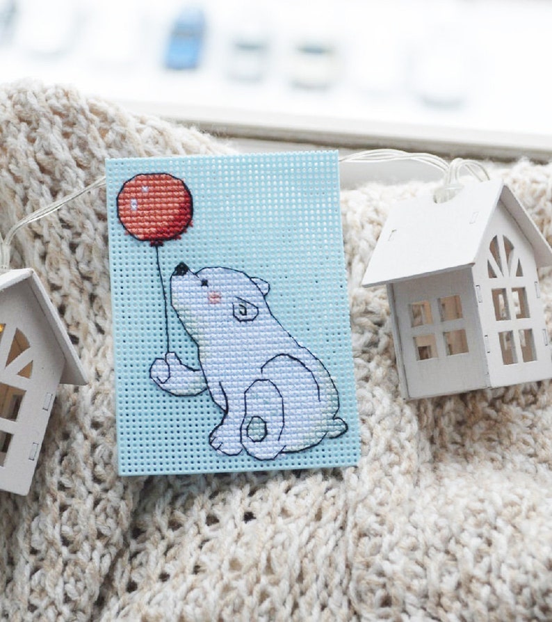 May include: A cross-stitch art piece featuring a white polar bear holding a red balloon against a light blue background. The bear has pink cheeks and is stitched on a square of perforated canvas. Two small white house-shaped decorations are in the background.