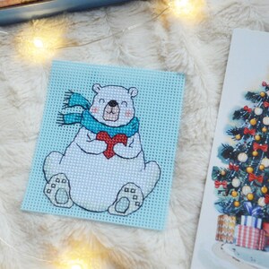 May include: A cross-stitch design of a polar bear holding a red heart, wearing a blue scarf, on a light blue background. A Christmas tree illustration is visible on the right. The background is a fluffy, white surface.