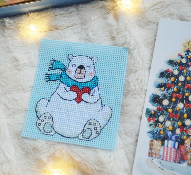 May include: A cross-stitch design of a polar bear holding a red heart, wearing a blue scarf, on a light blue background. A Christmas tree illustration is visible on the right. The background is a fluffy, white surface.