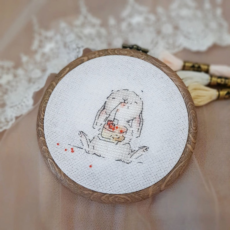 以下が含まれることがあります： 木製の輪の中に、白いウサギのクロスステッチ刺繍が施されています。ウサギは赤いフロスティングのケーキを持っています。