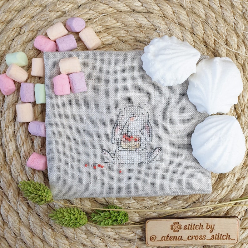 以下が含まれることがあります： ケーキを持ったウサギのクロスステッチパターン。下に「stitch by @alena_cross_stitch_」と書かれた木製の看板があります。