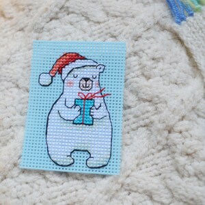 May include: A cross-stitch design of a polar bear wearing a Santa hat, holding a blue gift with a red bow. The bear is on a light blue background. The design is likely a Christmas ornament or decoration.