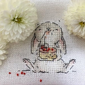 以下が含まれることがあります： 白いクロスステッチ刺繍で、赤いベリーが乗ったケーキを食べているウサギが描かれています。ウサギは白い背景に白い花に囲まれて座っています。