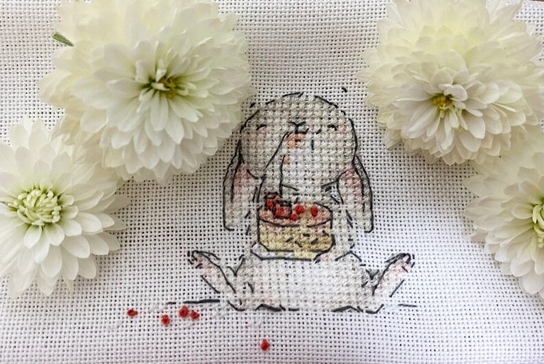 以下が含まれることがあります： 白いクロスステッチ刺繍で、赤いベリーが乗ったケーキを食べているウサギが描かれています。ウサギは白い背景に白い花に囲まれて座っています。