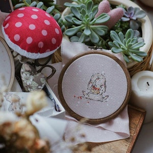 以下が含まれることがあります： 白い刺繍枠に、小さな動物がハートを持っているクロスステッチのデザインが施されています。枠はピンクの布地に置かれており、背景には赤と白のドット柄のキノコの針山があります。