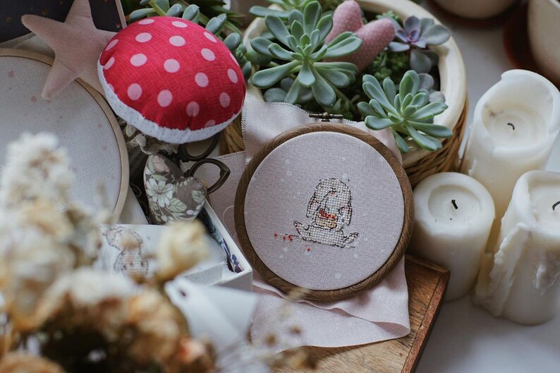 以下が含まれることがあります： 白い刺繍枠に、小さな動物がハートを持っているクロスステッチのデザインが施されています。枠はピンクの布地に置かれており、背景には赤と白のドット柄のキノコの針山があります。