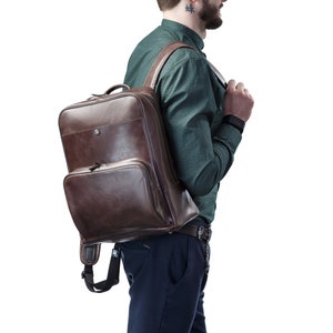 Könnte beinhalten: Ein Mann in einem grünen Hemd mit Knöpfen und blauen Hosen trägt einen braunen Lederrucksack. Er trägt eine Uhr am linken Handgelenk.