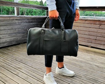 May include: A black leather duffel bag with a single zippered pocket on the front. The bag is being held by a person wearing black pants and white sneakers.