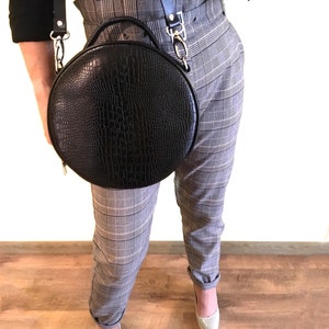 Può includere: Una borsa a tracolla rotonda nera in pelle con finitura coccodrillo. La borsa è indossata sopra una camicia nera e pantaloni grigi a quadri.