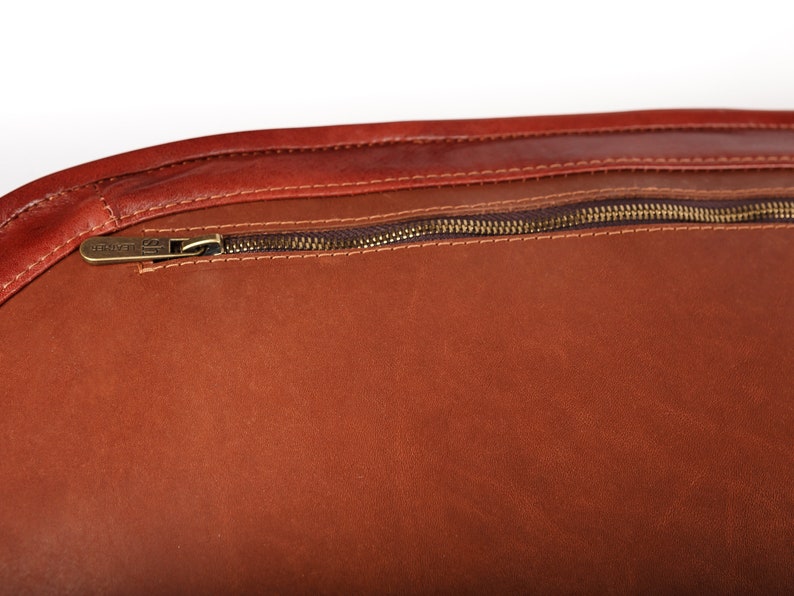 May include: Close-up of a brown leather bag with a zipper closure. The zipper has a brass pull with the word "UNIVERSAL" engraved on it.