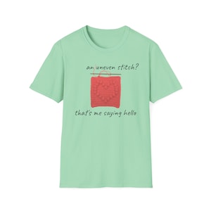 May include: A light green short-sleeve t-shirt featuring a graphic of a coral-colored square crochet piece with a heart pattern and a needle. The text above reads an uneven stitch? and the text below reads that's me saying hello.