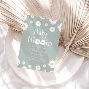 Lifestyle photo of the 'Baby in Bloom' daisy-themed invitation resting on a white plate beside a dried palm frond, styled on a white linen tablecloth.