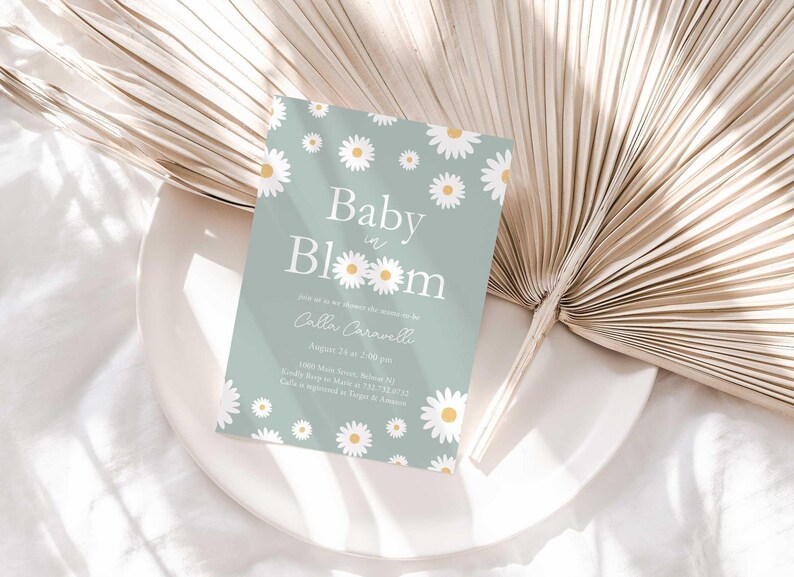 Lifestyle photo of the 'Baby in Bloom' daisy-themed invitation resting on a white plate beside a dried palm frond, styled on a white linen tablecloth.