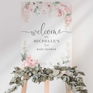 Styled photo of the "Welcome to Michelle's Baby Shower" sign on a wooden easel, with a lush eucalyptus greenery garland draped across the base of the easel.