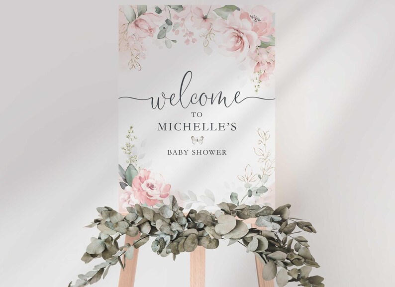 Styled photo of the "Welcome to Michelle's Baby Shower" sign on a wooden easel, with a lush eucalyptus greenery garland draped across the base of the easel.