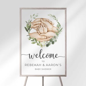 Welcome sign in a wood frame on an easel reading Welcome To Rebekah & Aaron's Baby Shower, with a watercolor illustration of parents' hands cradling baby feet inside a greenery wreath.