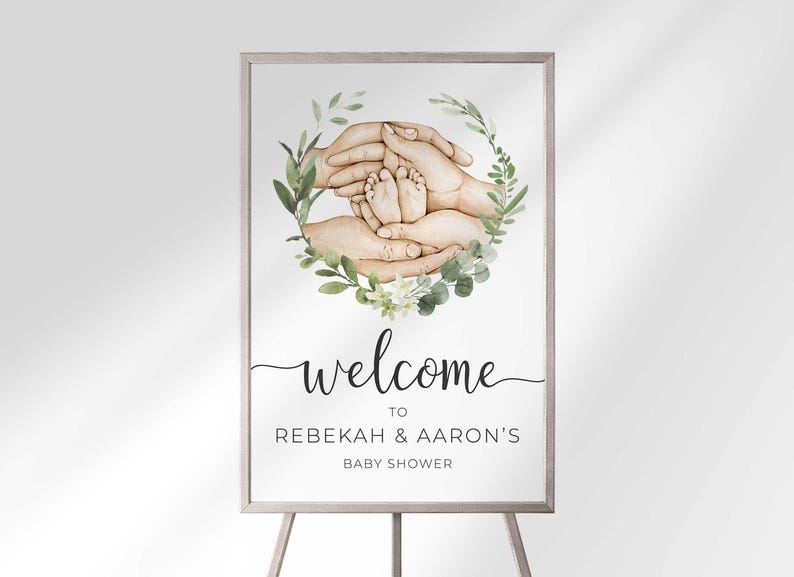 Welcome sign in a wood frame on an easel reading Welcome To Rebekah & Aaron's Baby Shower, with a watercolor illustration of parents' hands cradling baby feet inside a greenery wreath.