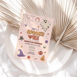 Olivia's Halloween-themed 'She's a Spooky One' first birthday invitation displayed on a white plate beside a dried palm frond, styled as a lifestyle photo.