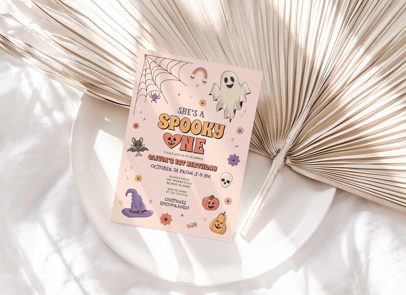 Olivia's Halloween-themed 'She's a Spooky One' first birthday invitation displayed on a white plate beside a dried palm frond, styled as a lifestyle photo.