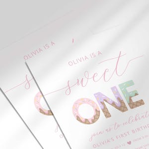 Angled close-up of two overlapping "Sweet ONE" invitations, highlighting the pastel confetti lettering and pink cursive script up close.