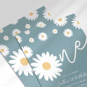Close-up angled shot of the 'ONE' daisy-themed invitation for Callie's 1st birthday in slate blue, showing the print quality and daisy pattern detail.