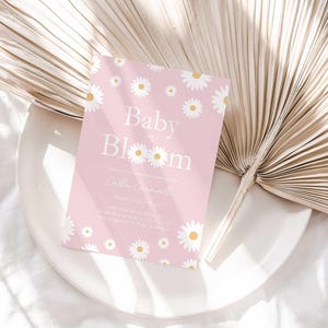 Lifestyle photo of the 'Baby in Bloom' daisy-themed invitation resting on a white plate beside a dried palm frond, styled on a white linen tablecloth.