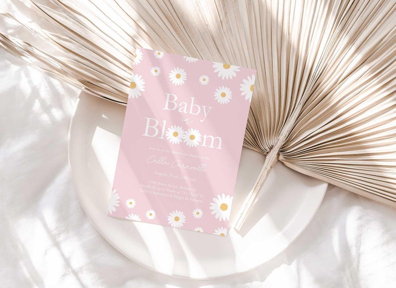 Lifestyle photo of the 'Baby in Bloom' daisy-themed invitation resting on a white plate beside a dried palm frond, styled on a white linen tablecloth.