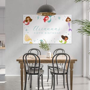 May include: A rectangular canvas print featuring mermaids and the text "Arianna's Fifth Birthday." The mermaids have various hair and tail colors, with a light blue background and star accents. The print is displayed above a wooden dining table.