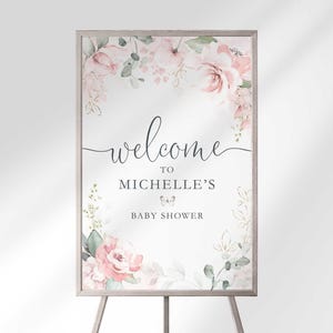 "Welcome to Michelle's Baby Shower" sign displayed on a wooden easel, with blush pink and white floral bouquets and greenery framing the top and bottom corners.
