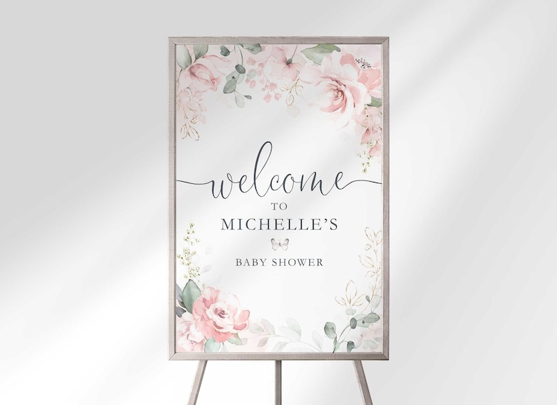"Welcome to Michelle's Baby Shower" sign displayed on a wooden easel, with blush pink and white floral bouquets and greenery framing the top and bottom corners.