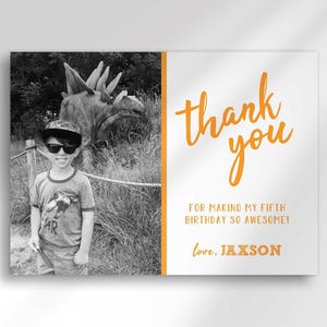 Full close-up view of the photo thank you card, showing a black-and-white photo of a boy wearing sunglasses and a cap at a dinosaur exhibit, and orange "thank you" text reading "for making my fifth birthday so awesome! love, Jaxson."