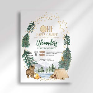 Camping-themed first birthday invitation with illustrated pine trees, a bear, a tent, and a campfire scene framed by a gold star arch, reading "One Happy Camper, hike on over to celebrate Alexander's first birthday" with the party date, time, and location.