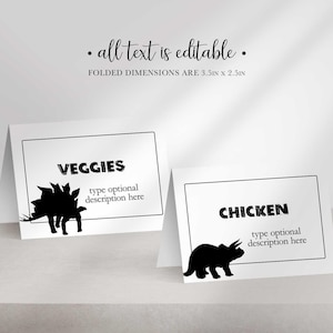 Two folded place cards stand side by side, one reading VEGGIES with a stegosaurus silhouette and the other CHICKEN with a triceratops silhouette.