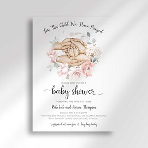 以下が含まれることがあります： 「For This Child We Have Prayed」のテキストが入ったベビーシャワーの招待状。ピンクの花に囲まれた、赤ちゃんの足を抱える手の水彩イラストが特徴です。招待状には、両親の名前が記載されています。
