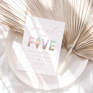 Styled lifestyle photo of the invitation propped on a white plate against a gold dried palm frond, on a white linen surface.