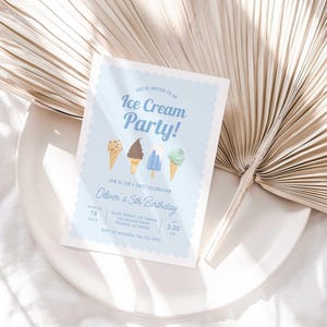 Styled lifestyle photo of the light blue ice cream invitation propped on a white plate against a gold dried palm frond, on a white linen surface.