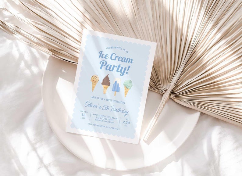 Styled lifestyle photo of the light blue ice cream invitation propped on a white plate against a gold dried palm frond, on a white linen surface.