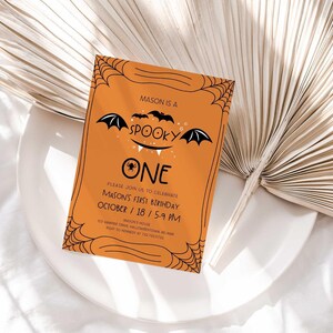 Mason's Halloween-themed 'Spooky One' first birthday invitation displayed on a white plate beside a dried palm frond, styled as a lifestyle photo.